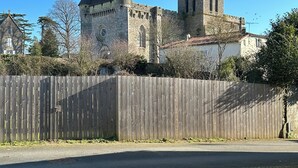 Een historisch gebouw van steen met een opvallende toren en boogvensters, omgeven door een houten hek en bomen.
