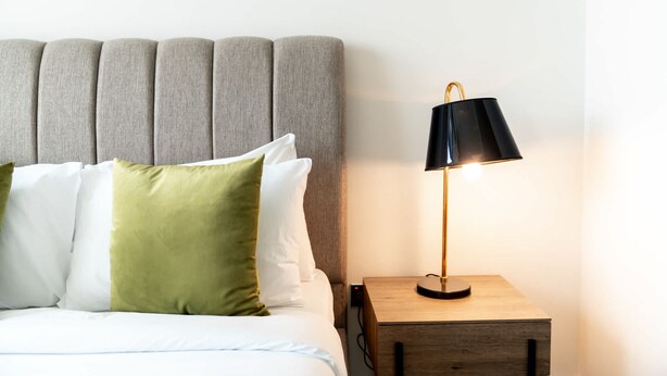 A neatly made bed with a grey upholstered headboard, a bedside table with a lamp, and a wooden nightstand. - Free Housekeeping Visits During Your Stay - Stayshort - Modern 2BR Retreat in Dubai South for 6 (Dubai)