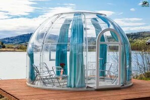 Une structure en forme de dôme transparent, dotée de rideaux, d’une terrasse en bois et de chaises.
