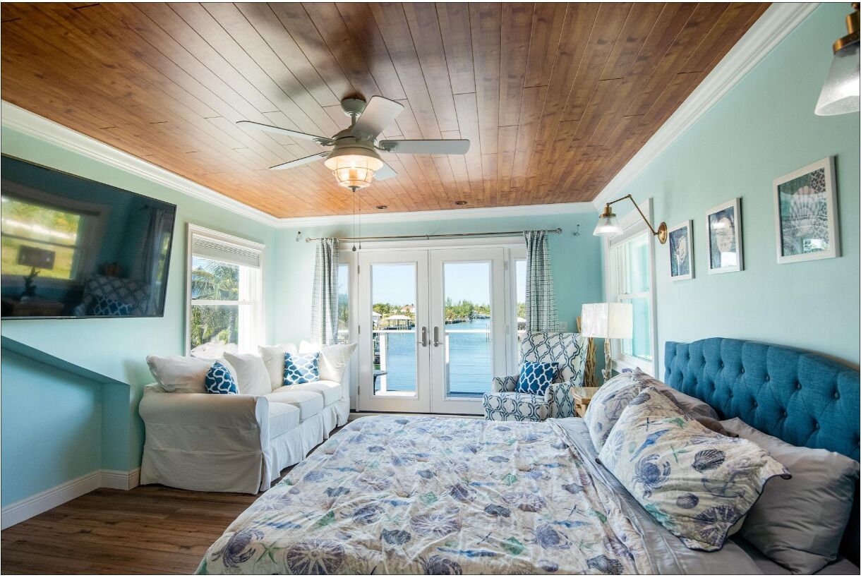 A bedroom with a large bed, a ceiling fan, a view of the outdoors through a sliding door, and a window with curtains.