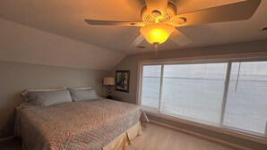Une chambre à coucher comprenant un lit, un ventilateur de plafond, une fenêtre avec des stores et une table de chevet avec une lampe et un cadre photo.