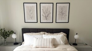 Ett sovrum med en säng, sängbord, ett litet bord och inramade botaniska illustrationer på väggen.