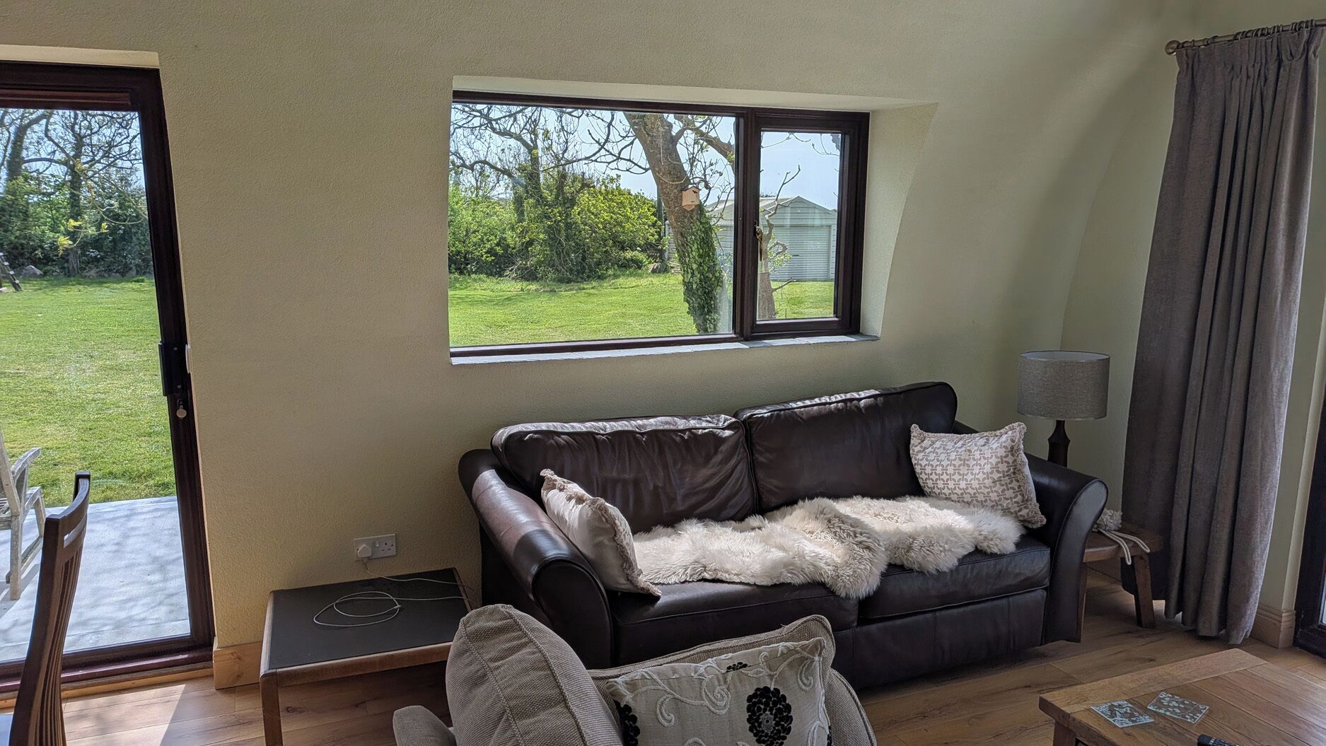 Ein Wohnzimmer mit einer Ledersofa, einem kleinen Tisch, einer Lampe und einem Fenster mit Blick auf einen Garten.
