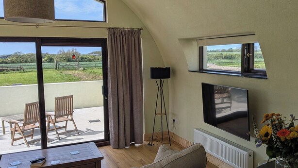 A living room with a TV, sofa, and a view of a green landscape through a sliding door. - The Rookery (Tacumshane)