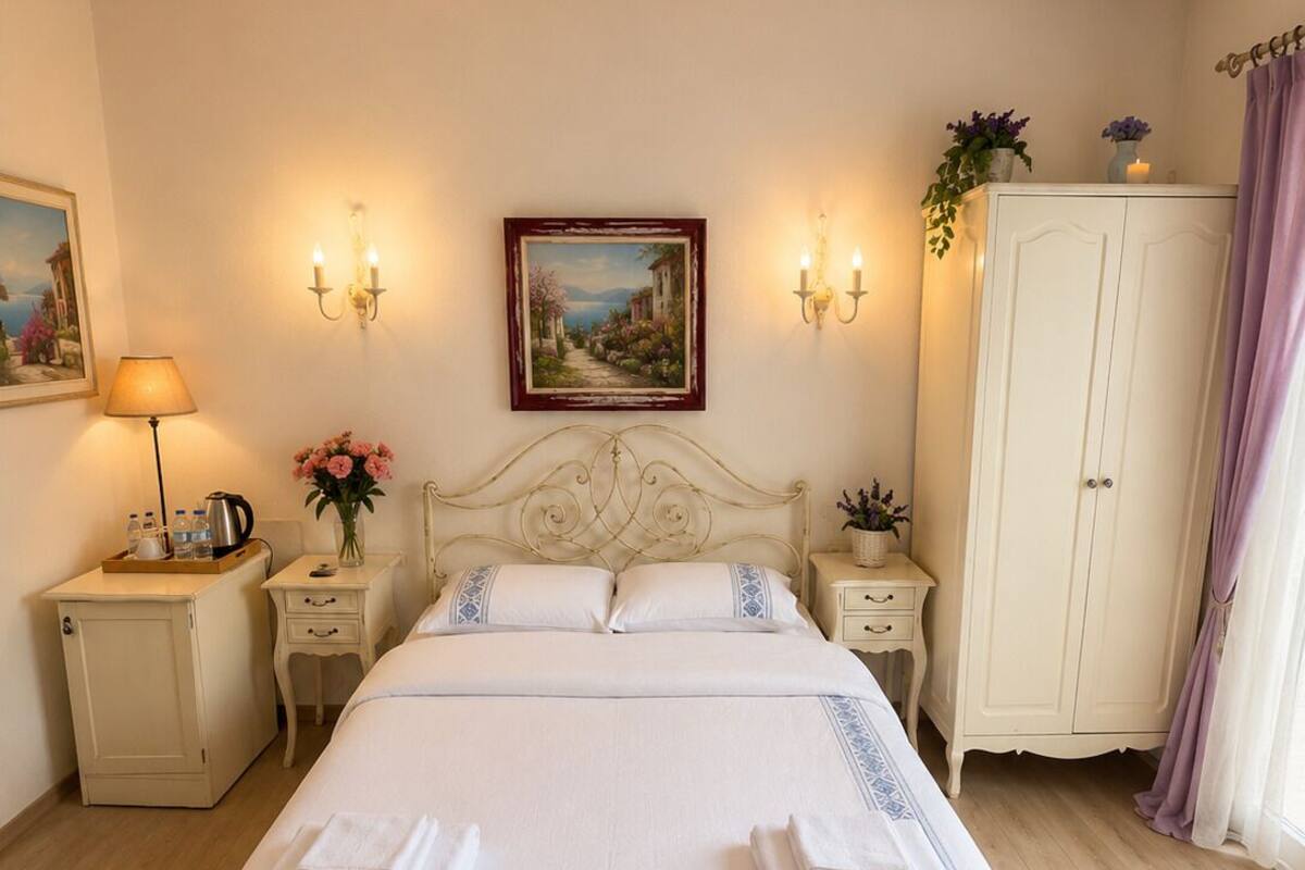 Una cama bien hecha con sábanas blancas y detalles azules, una mesita de noche con una lámpara, una cómoda con un florero y un cuadro en la pared.