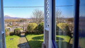 A view from a window overlooking a garden with a stone pathway, a white lattice structure, and a distant landscape with houses and mountains. - Garth Celyn (Porthmadog)