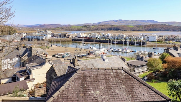 A coastal marina with boats, residential buildings, and a view of the sea and hills in the background. - Garth Celyn (Porthmadog)