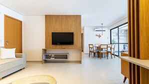 En moderne stue med et fladskærms-tv monteret på en træpanel, en grå sofa, et spisebord med stole og et stort vindue, der giver naturligt lys.
