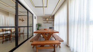 Et moderne spiseområde med et træbord og bænke, et køkken i baggrunden og store vinduer med lette gardiner.