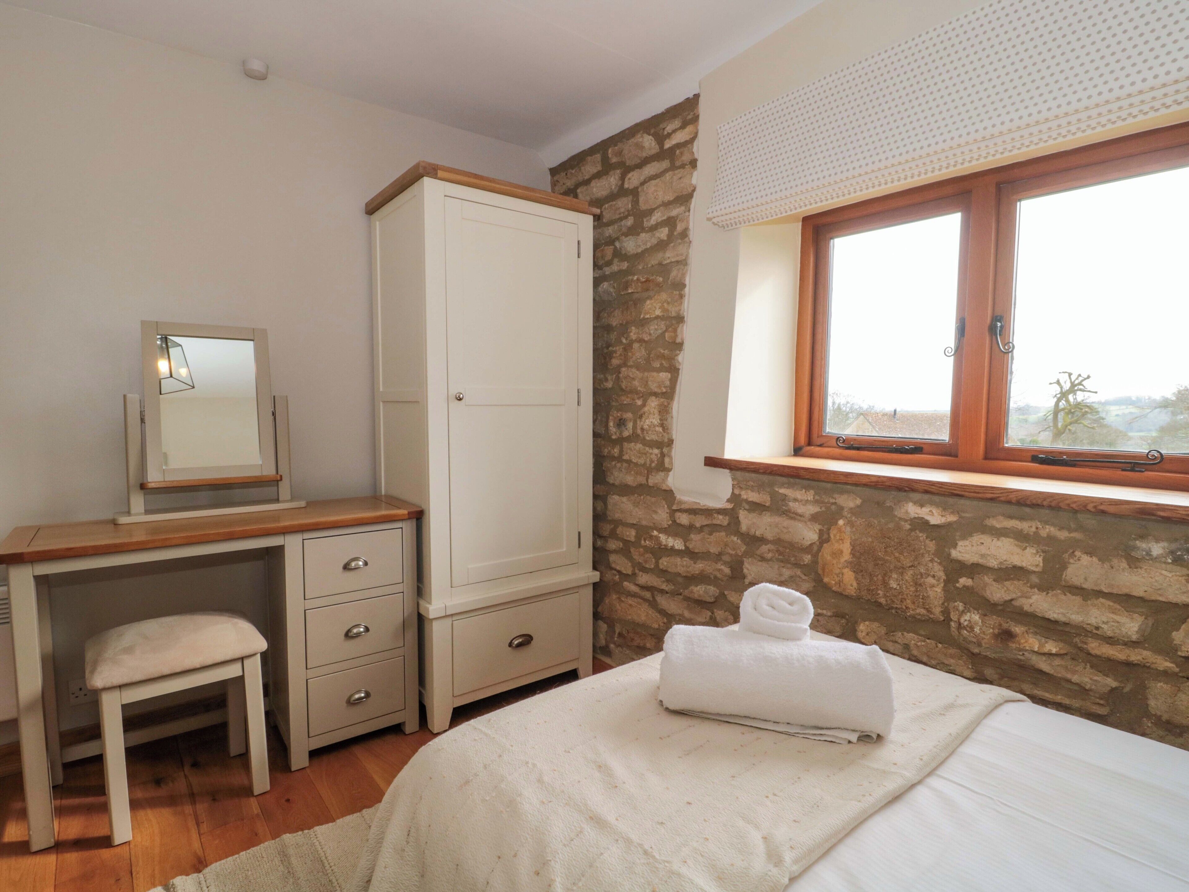 A bedroom with a wooden desk, a white wardrobe, a small stool, a bed with a white blanket, and a window with a view.
