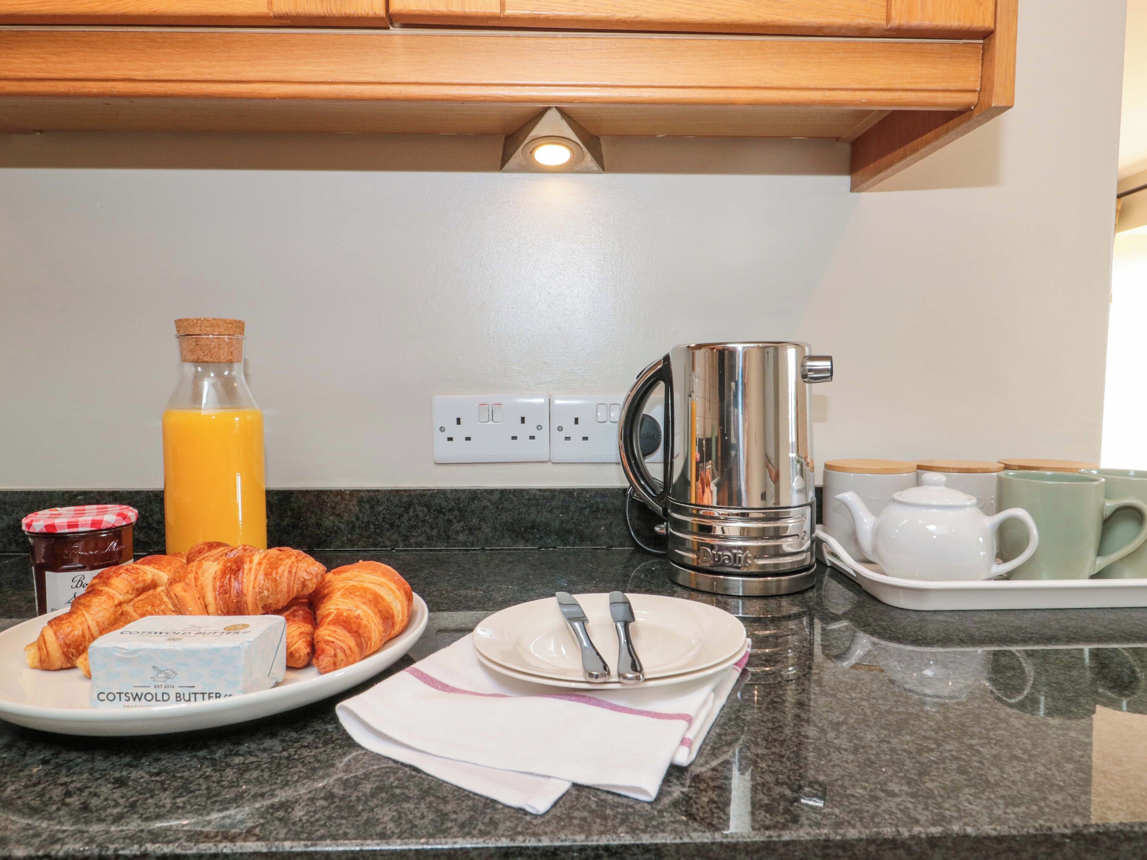 A breakfast setup with croissants, butter, a bottle of orange juice, a kettle, and teapots on a kitchen counter.
