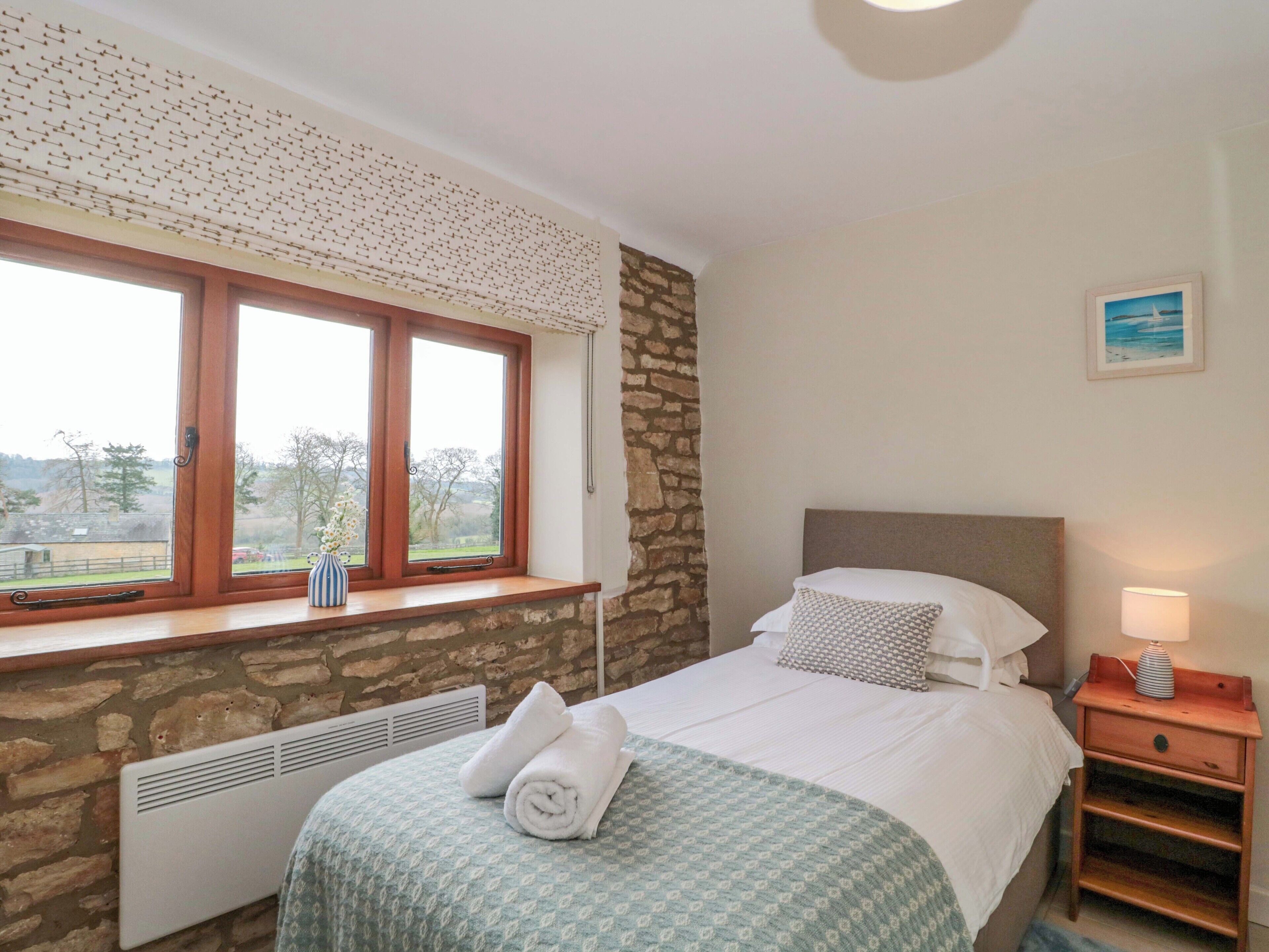 A bedroom with a stone wall, a bed with a patterned bedspread, a bedside table with a lamp, and a window with a view of the outdoors.