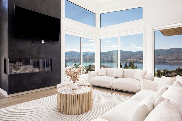 Ein modernes Wohnzimmer mit einem großen Flachbildfernseher, einem Kamin, einem weißen Sofa und Blick auf einen See und Berge durch große Fenster.