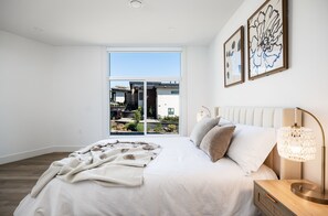 Ein Schlafzimmer mit einem großen Bett, einem Nachttisch, einer Lampe und Blick auf ein Gebäude durch eine Glastür.