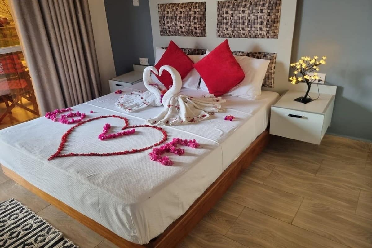 A neatly made bed with a heart-shaped decoration and swan-shaped towels, a bedside table with a lamp, and a woven headboard.