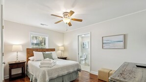 A bedroom with a wooden floor, a bed with a headboard, two bedside tables with lamps, a ceiling fan, a window with blinds, a framed picture on the wall, and a door leading to a bathroom. - Camp Carolina 3BR 2BA home with private pool close to beach! (Isle Of Palms)
