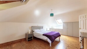 A bedroom with a sloped ceiling, a bed with a white headboard, a nightstand with a lamp, a wardrobe, and a skylight.