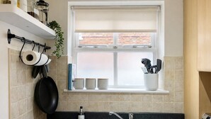 A kitchen with a sink, a window with a blind, a hanging pot rack, and a shelf with cups and utensils. - 3BR Jericho Oxford Home | Walk To The University (Oxfordshire)