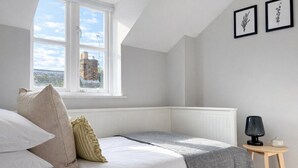 A bedroom with a large window, a bed with pillows, a bedside table with a lamp, and framed pictures on the wall. - 3BR Jericho Oxford Home | Walk To The University (Oxfordshire)