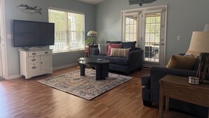 Una sala de estar con televisor de pantalla plana, ventilador de techo, sofá, mesa de centro, alfombra y ventana con persianas.