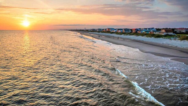 Una playa con un atardecer, aguas tranquilas y casas a lo largo de la costa.
