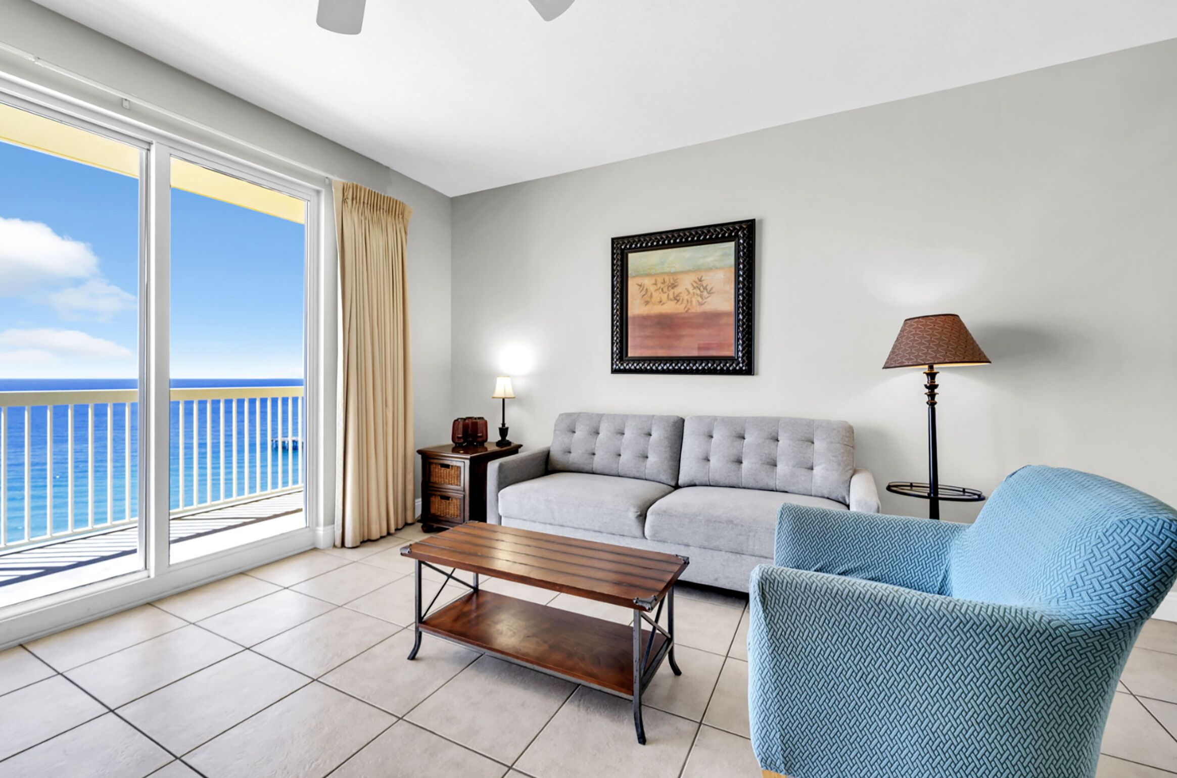 A living room with a sofa, armchair, coffee table, and a lamp. There is a painting on the wall and a window with a view of the ocean.