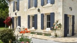 A stone building with multiple windows and shutters, a patio with outdoor seating, and a garden with flowers. - In the Perrines garden, in the heart of Aunis (Marsais)