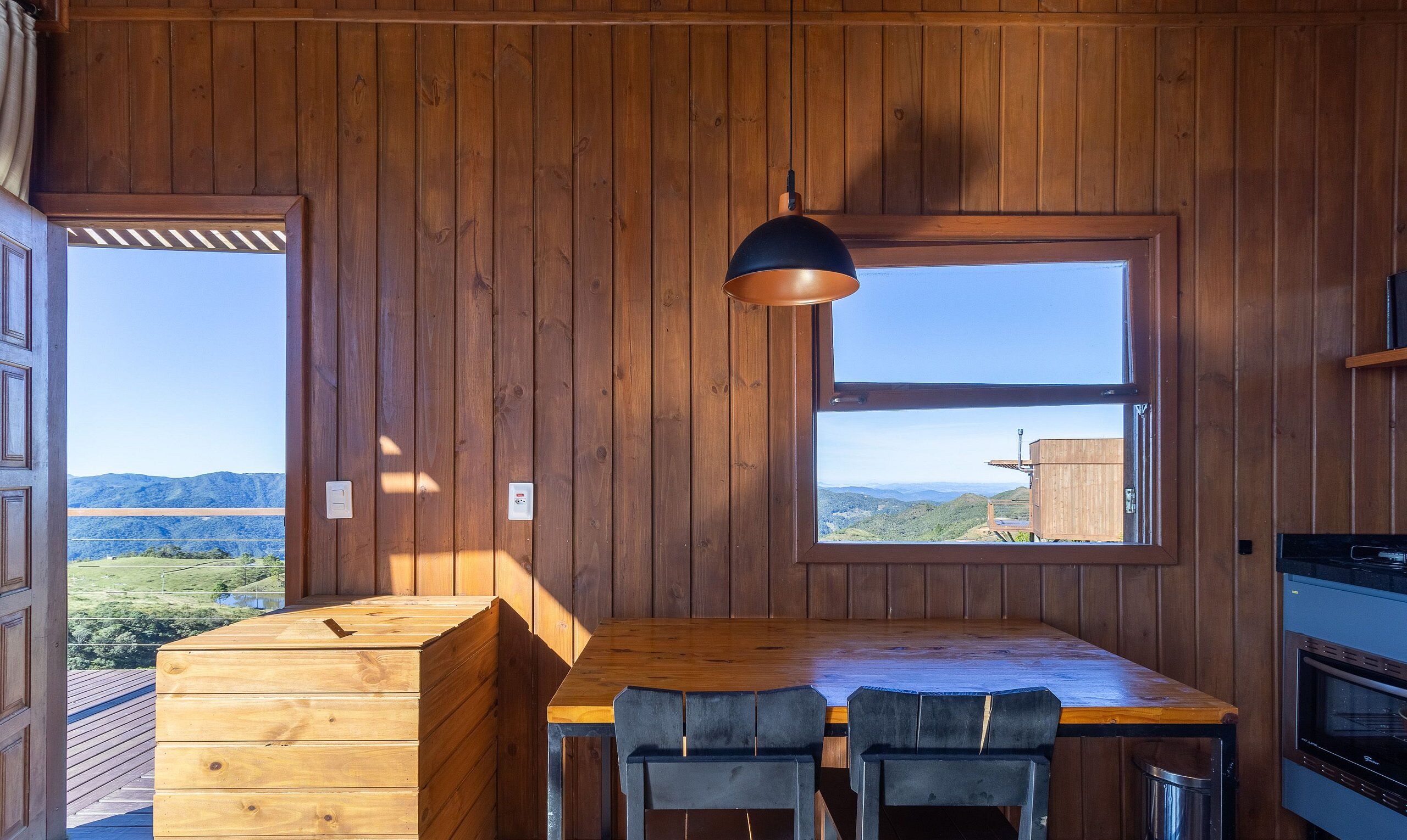 A wooden interior with a dining table and chairs, a window with a view, and a hanging light fixture.