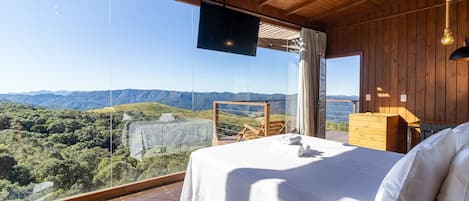 A bedroom with a large bed, wooden ceiling, and a TV. It has a view of mountains and greenery.