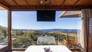 A bedroom with a large window overlooking a scenic landscape, a bed, and a wooden ceiling.