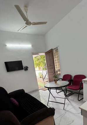 A room with a ceiling fan, a flat-screen TV mounted on the wall, a small table with a vase, and two red chairs. - Little Chandhana Guest House (Kottayam)
