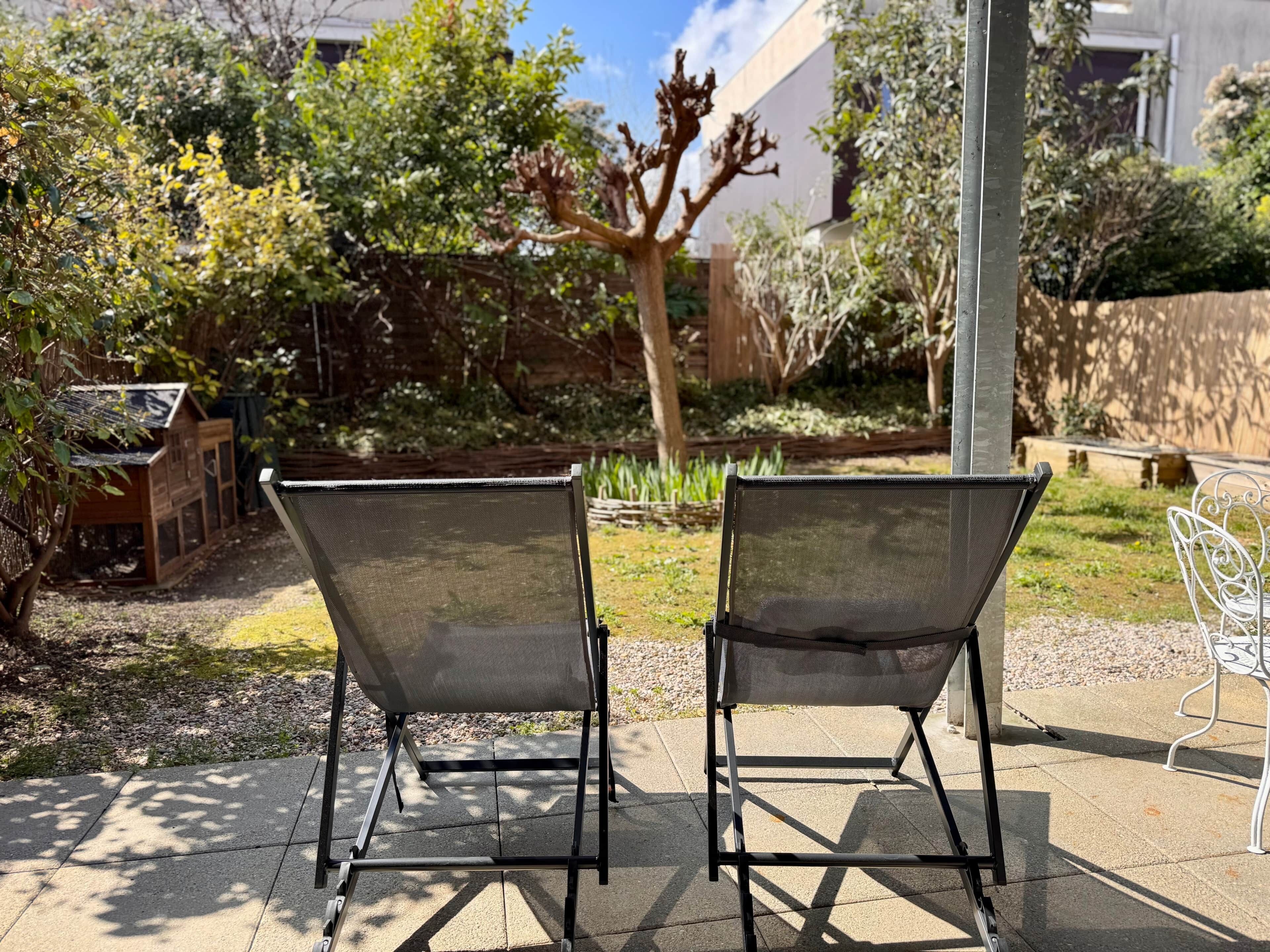 Due sedie da esterno con schienale a rete e struttura in metallo, poste su un patio lastricato con un giardino e una struttura in legno sullo sfondo.