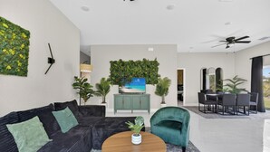 Una sala moderna con un sofá negro, un sillón verde, una mesa de centro de madera, un televisor, un armario verde y una zona de comedor con una mesa y sillas.