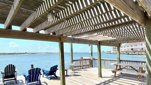 A wooden deck with chairs overlooking a body of water, a covered area with a view of a distant building. - Spacious • Walk to Beach • Pool & Lake Padre Views • Fishing Dock [6 night min.] (Corpus Christi)