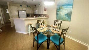 A dining area with a glass table and chairs, a kitchen in the background, and a painting of a beach scene on the wall. - Spacious • Walk to Beach • Pool & Lake Padre Views • Fishing Dock [6 night min.] (Corpus Christi)