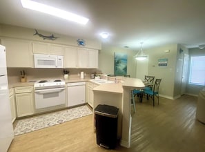 A compact kitchen with white cabinets, a microwave, stove, and dishwasher. There's a small trash bin and a dining area with chairs and a table. - Spacious • Walk to Beach • Pool & Lake Padre Views • Fishing Dock [6 night min.] (Corpus Christi)