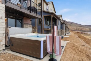 Un bain à remous à l’extérieur d’une maison à deux étages, dotée d’une façade en pierre, d’un balcon et offrant une vue sur un paysage vallonné.