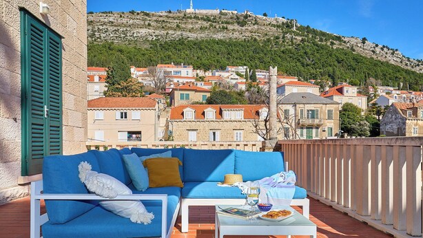 A terrace with a blue sofa, white coffee table, and a view of a hillside town. - Green Horizon - One Bedroom Apartment, Sleeps 4 (Dubrovnik)