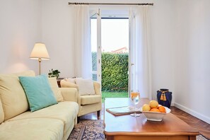 Ein Wohnzimmer mit einer Couch, einem Sessel, einem Couchtisch und Blick auf einen grünen Hecken durch eine Glastür.