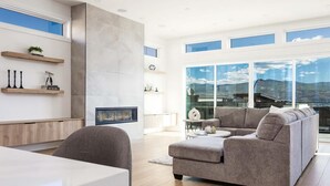 Een moderne woonkamer met een grote modulaire bank, een eettafel en een open haard. Er zijn houten planken met decoratieve objecten en een groot raam met uitzicht op de bergen.