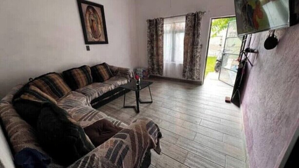 A living room with a sectional sofa, a coffee table, a flat-screen TV mounted on the wall, and a window with curtains.