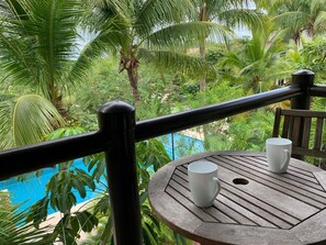 Ein Holztisch mit zwei weißen Tassen auf einem Balkon mit Blick auf einen Pool, umgeben von üppiger Vegetation.