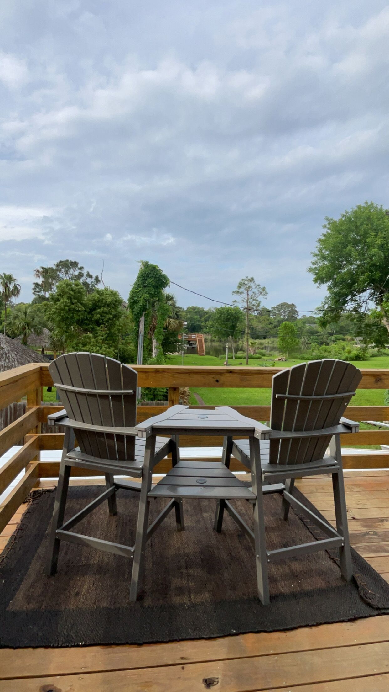 Eine Terrasse mit zwei Adirondack-Stühlen und einem kleinen Tisch, mit Blick auf eine Rasenfläche mit Bäumen und ein entferntes Gebäude.