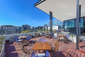 A rooftop terrace with wooden tables and blue chairs, overlooking a cityscape.
