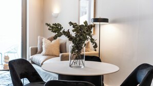 Ein modernes Wohnzimmer mit einem runden Tisch, zwei schwarzen Stühlen, einer Glaskaraffe mit Grünpflanzen, einem Sofa mit beigen Kissen und einer Stehlampe.