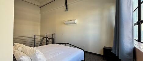 A single bed with a metal frame and white bedding in a room with a plain white wall, a ceiling light, and an air conditioning unit.
