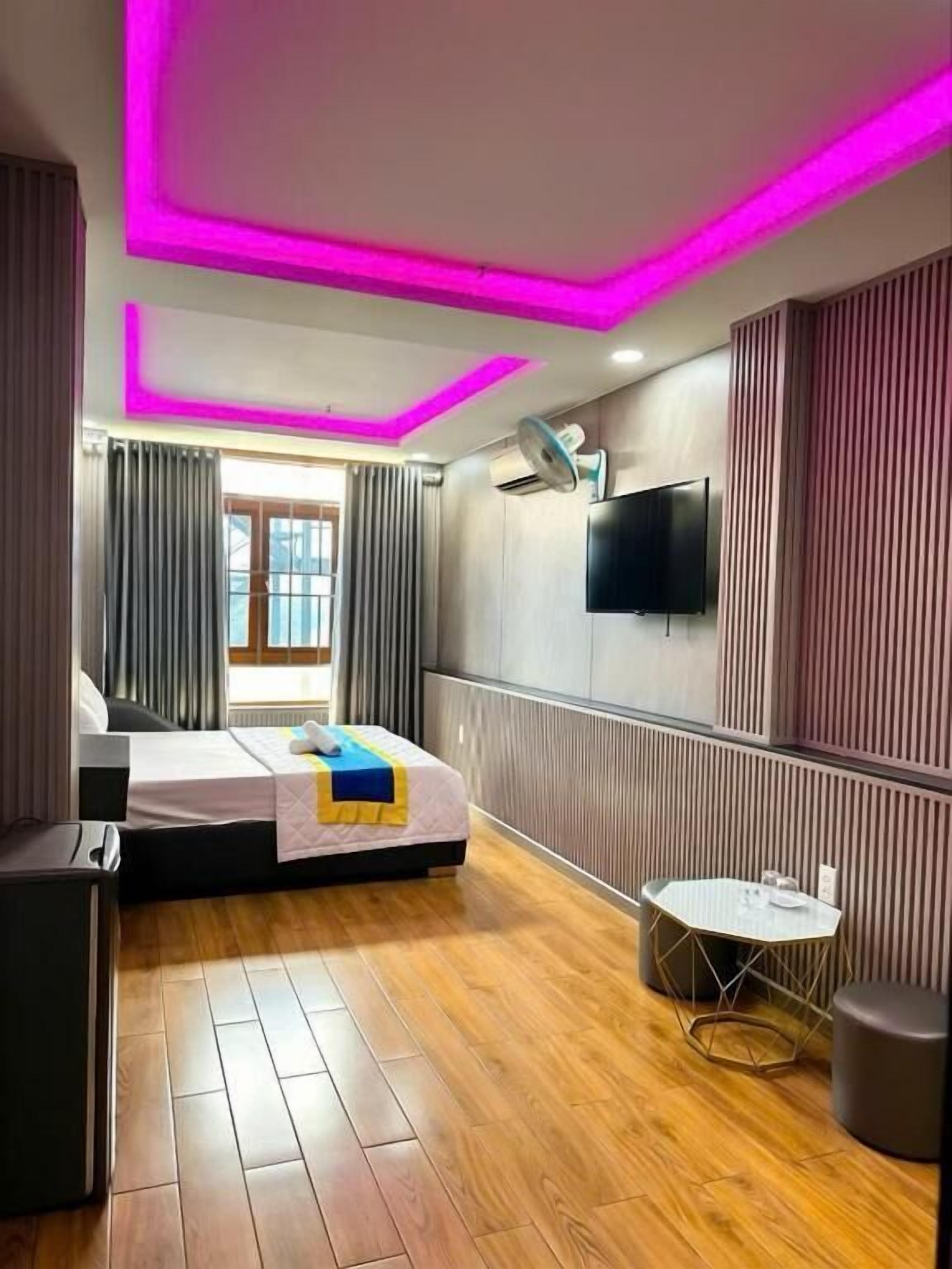 A hotel room with a bed, bedside table, a small round table, a flat-screen TV mounted on the wall, and a window with curtains.