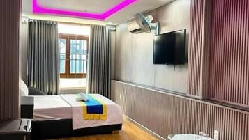 A hotel room with a bed, bedside table, a small round table, a flat-screen TV mounted on the wall, and a window with curtains.