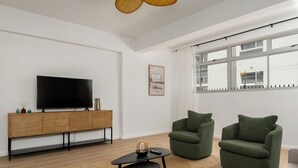Un salon moderne avec un téléviseur à écran plat, un meuble en bois, deux fauteuils verts, une petite table basse noire et une grande fenêtre donnant sur un immeuble.