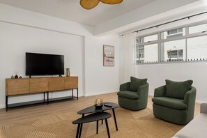 Ein modernes Wohnzimmer mit Fernseher, Holzschrank, zwei grünen Sesseln, einem kleinen schwarzen Couchtisch und einem großen Fenster mit Blick auf ein Gebäude.